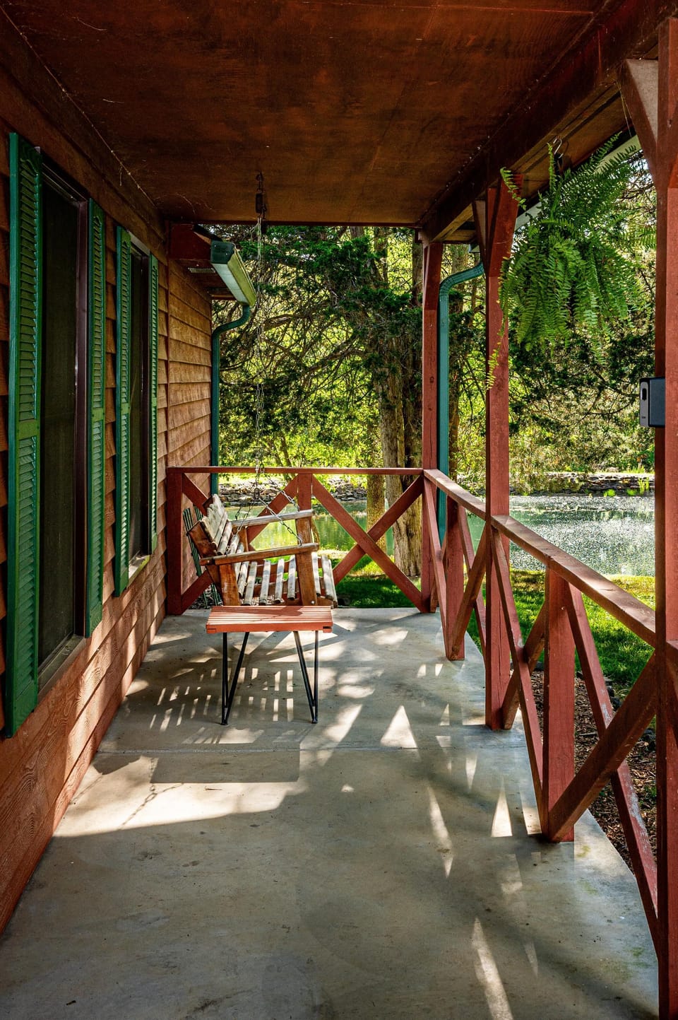 Wooden porch swing and small table sit on a covered concrete porch overlooking a pond and trees.