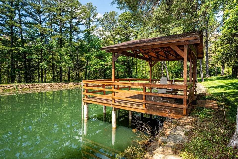 Covered wooden dock with seating extends over a green pond, surrounded by trees and stone edging.