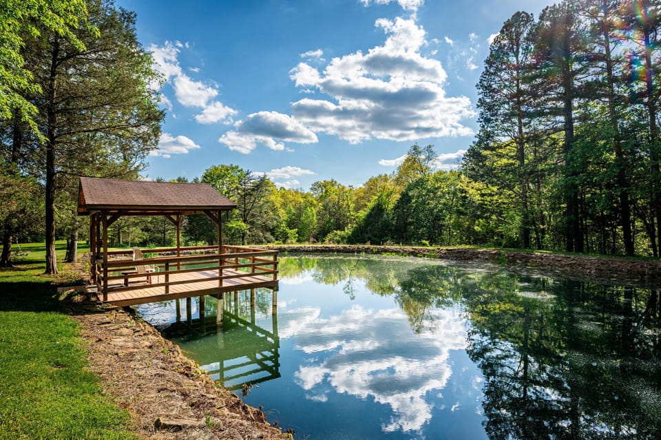 Covered wooden dock with Adirondack chairs extends over a stone-edged pond surrounded by trees and grassy lawn.