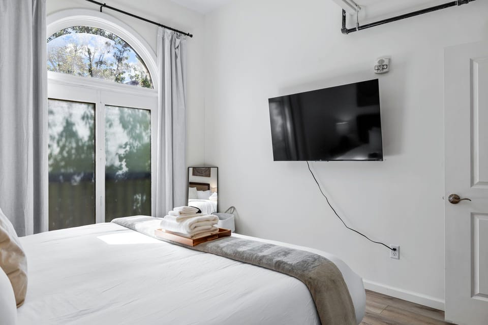 Bedroom view showing the bed, wall-mounted TV, doorway, and natural light.