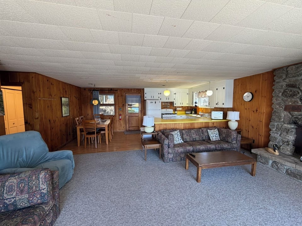Unwind in your cozy open-plan retreat with rustic wood paneling, stone fireplace, and fully equipped kitchen perfect for family gatherings.