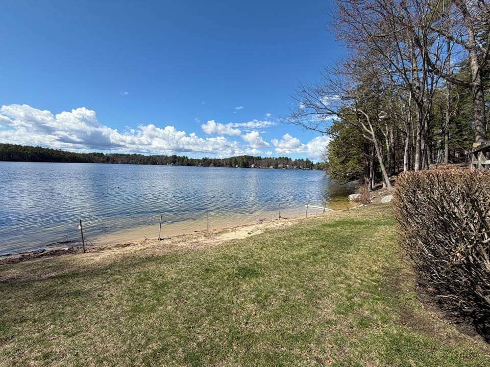 Pristine lakefront with private beach area and manicured lawn, surrounded by mature trees and distant shoreline homes.