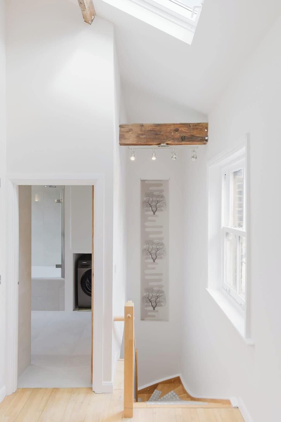 Capture the upper stair landing area illuminated by a roof skylight, showing the top of the staircase, exposed timber beams, and an doorway opening into the laundry bathroom.