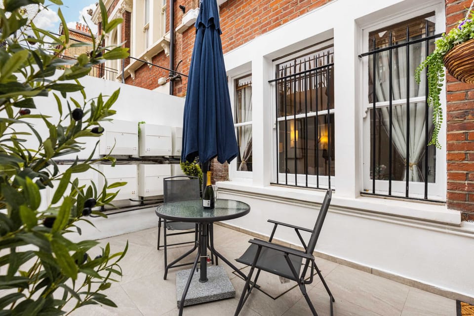 Look across the tiled courtyard seating area toward the potted olive tree, navy patio umbrella, and lower-level building facade.