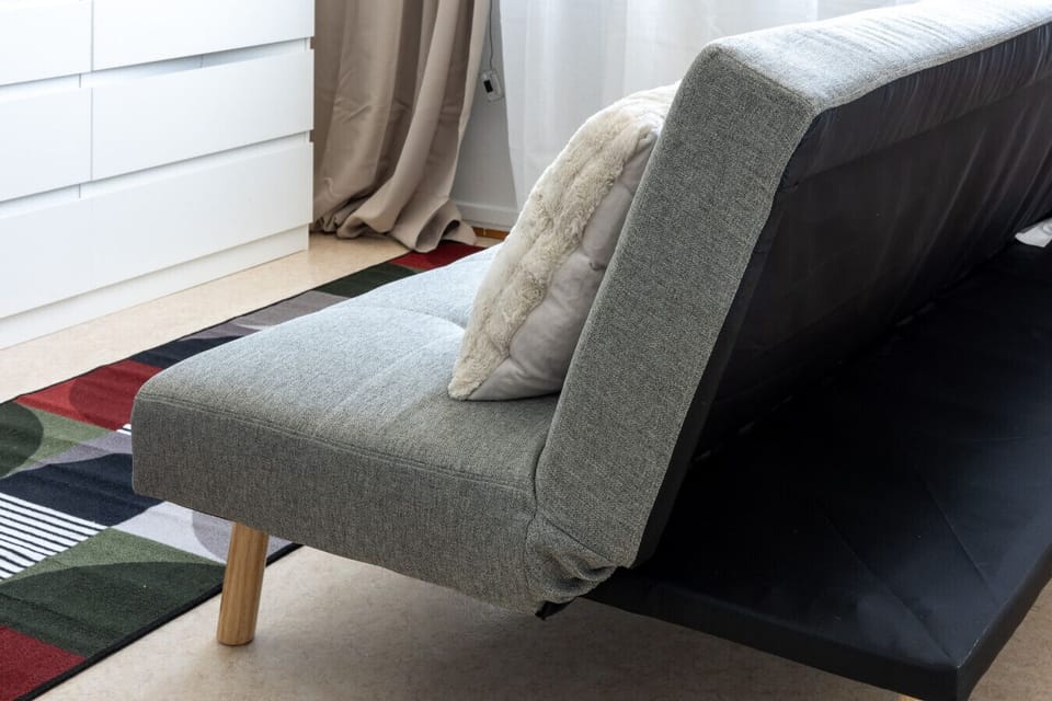 Close-up of a contemporary grey fabric sofa bed featuring a plush cream pillow, positioned on a patterned rug next to white drawers.