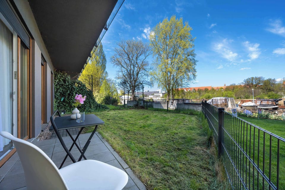 A small black table and two chairs are positioned in the garden, overlooking the green lawn. The surrounding fence provides a sense of privacy.