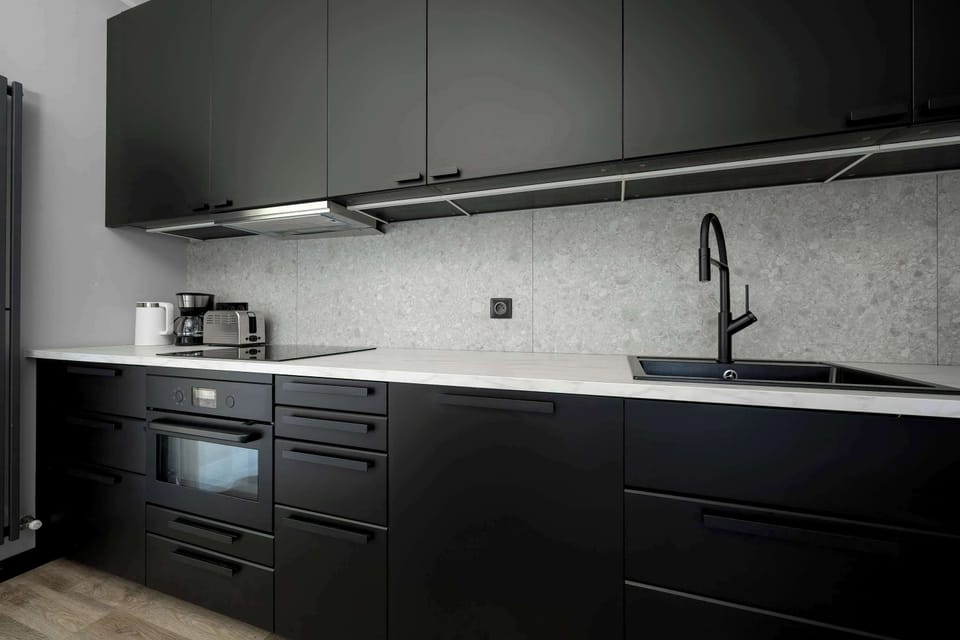 The kitchenette is designed in a sleek black finish with modern appliances including a built-in oven and induction hob. A dark backsplash with a subtle pattern adds a sophisticated touch.