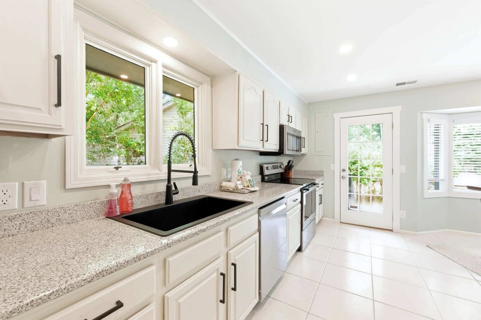 Granite countertops and crisp white cabinetry create a clean, airy kitchen workspace. Afternoon glow pours through the broad window, framing a view of lush garden greenery while you prepare your favorite morning coffee.