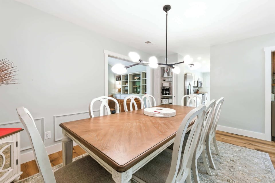 Warm wood grains and elegant white seating anchor this airy dining space, illuminated by a sculptural modern chandelier. Soft gray walls and open sightlines create a calm, social atmosphere perfect for long morning coffees and shared meals.