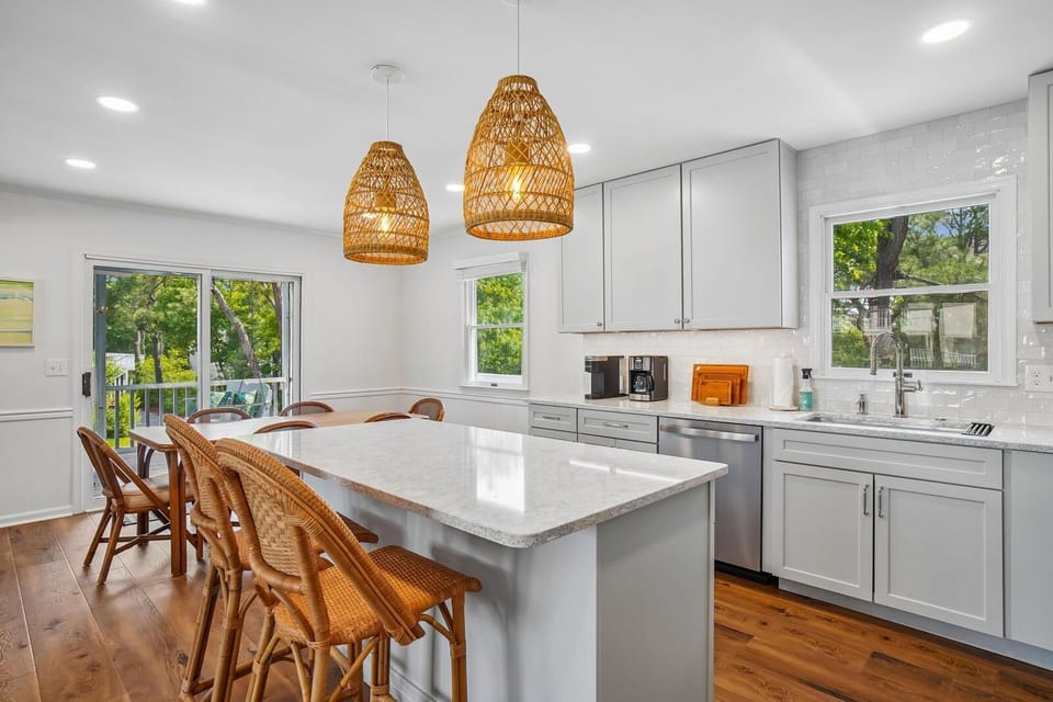 Gather around this bright kitchen island with friends and family. Natural light pours in, woven pendants glow warmly overhead, and your morning coffee station awaits on gleaming counters.