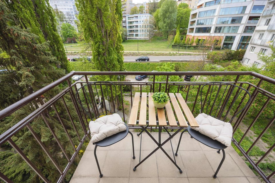 A balcony with a small outdoor table and two chairs, surrounded by greenery. It’s a pleasant spot for morning coffee with a view of the courtyard.