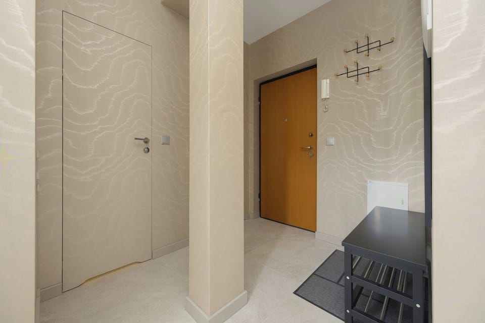 A clean and minimal hallway with light-colored walls and a wooden entrance door. A wall-mounted coat rack and a small bench provide practical storage and seating.