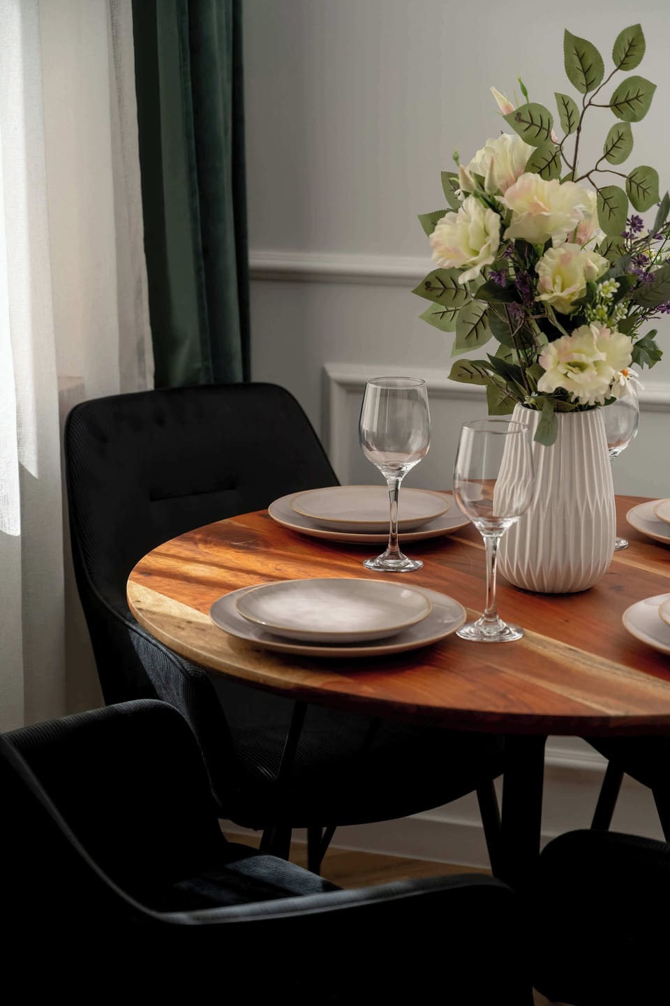 A cozy dining corner with a round table set with plates and glassware, surrounded by dark upholstered chairs. A vase with fresh flowers adds a decorative touch, while a window with curtains brings in natural light. 