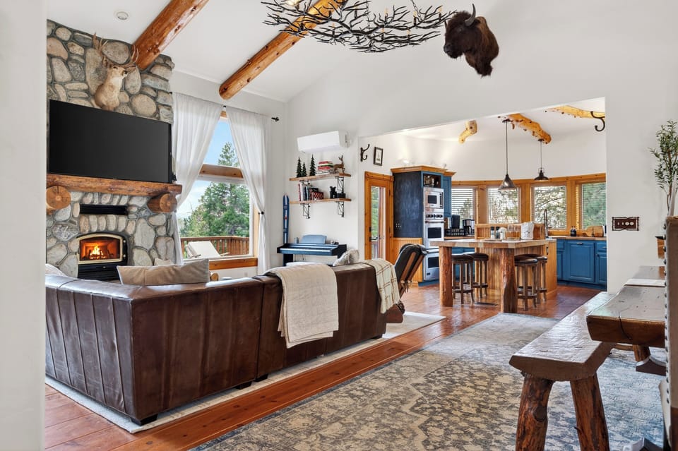 Warm cabin‑style living room with a stone fireplace, leather seating, and forest views through large windows.