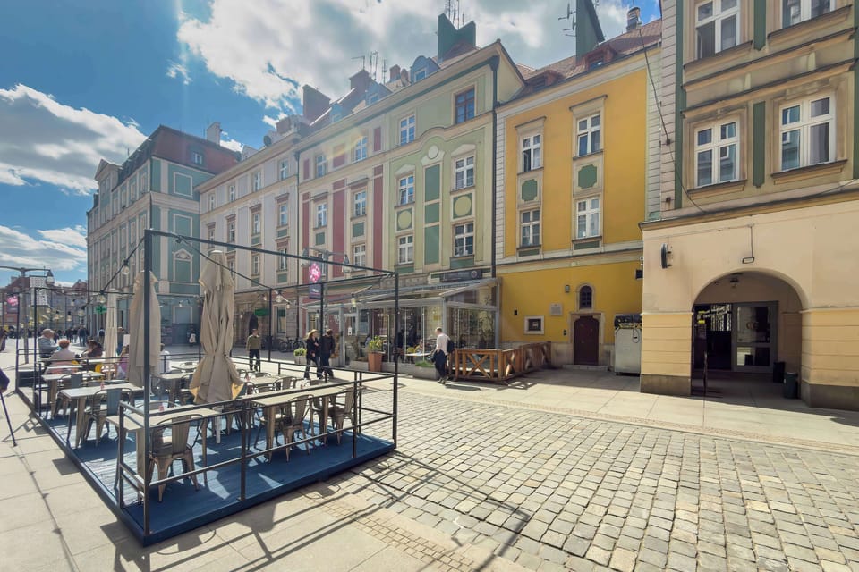 An outdoor view of a charming city square with historic buildings and café seating. The atmosphere is lively and full of urban character.