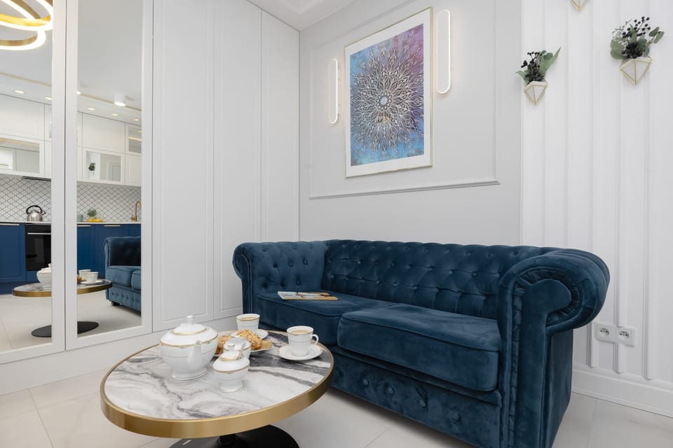 A luxurious navy blue tufted sofa sits against a white paneled wall decorated with a subtle piece of art. In front of it, a round marble coffee table with gold accents holds a small tea set, creating an elegant lounge area.