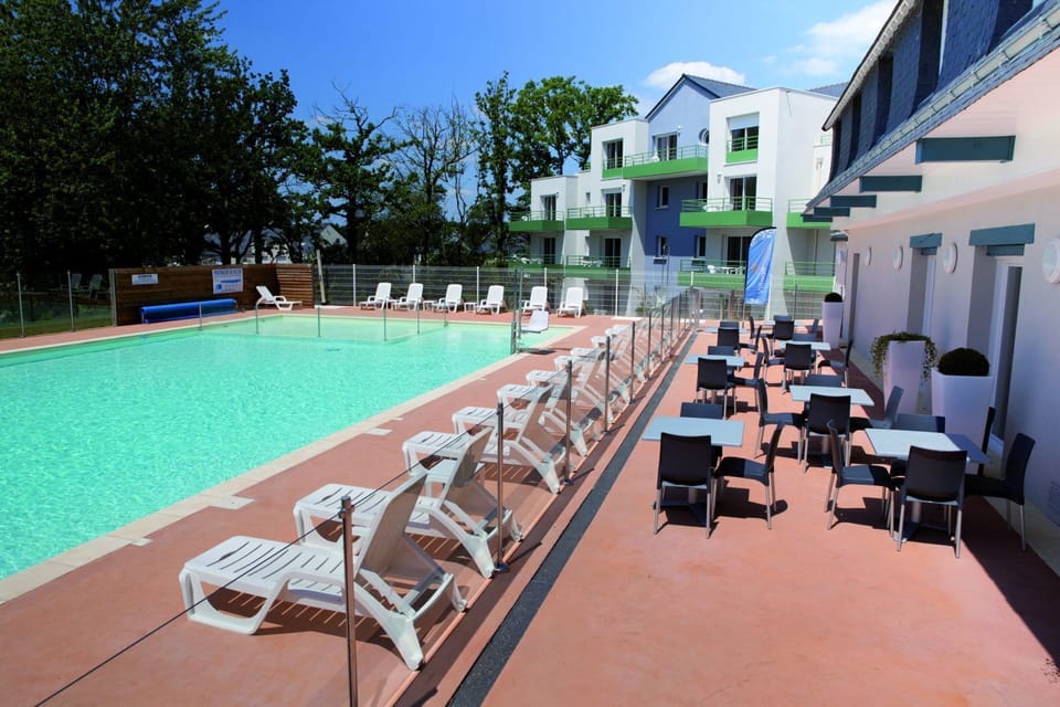 Pool terrace with white loungers on one side and outdoor café tables and chairs on the other, apartment building behind