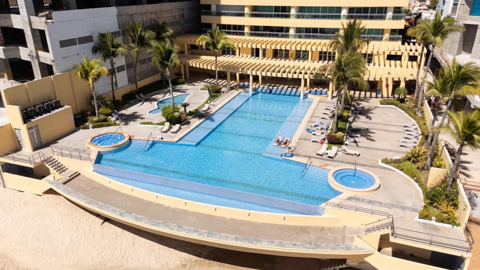 Infinity-edge pool with two circular hot tubs, palm trees, and a sandy beach below yellow pergolas.