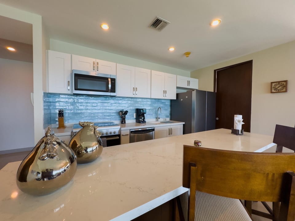 Quartz countertop island with wooden chairs overlooks white cabinetry and a glossy blue tile backsplash in the kitchen.