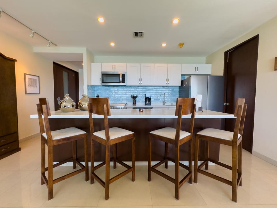 White quartz island with four wooden barstools faces a kitchen backsplash of glossy blue subway tiles.