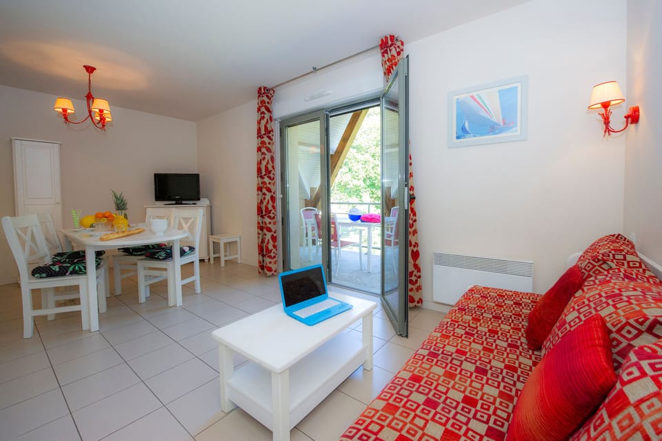 Open-plan living and dining with red sofa, wall-mounted TV, and glass doors opening to the furnished balcony