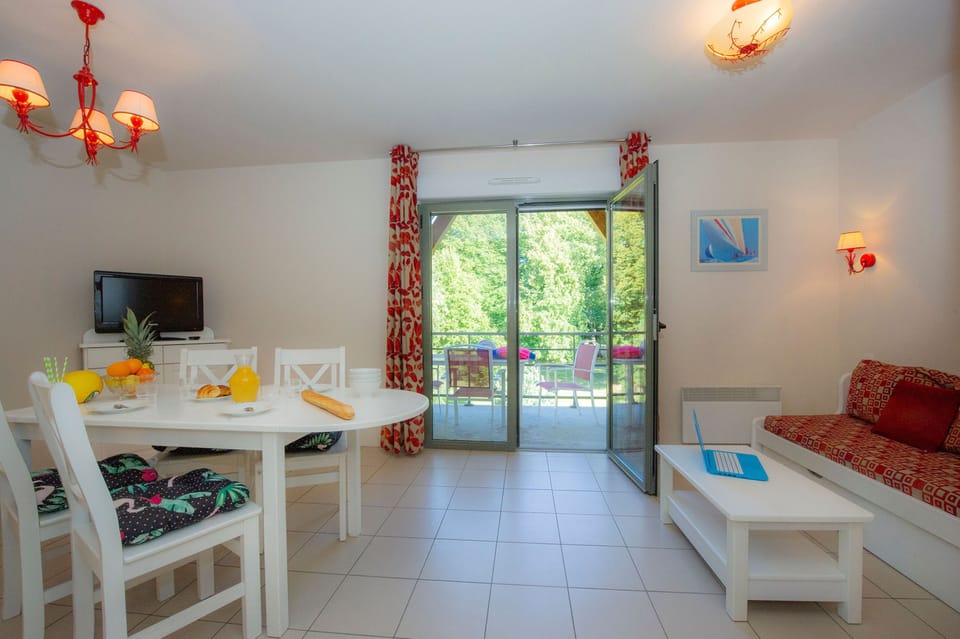 Open-plan living and dining area with round white table, four seats, wall-mounted TV, and sliding doors to the balcony
