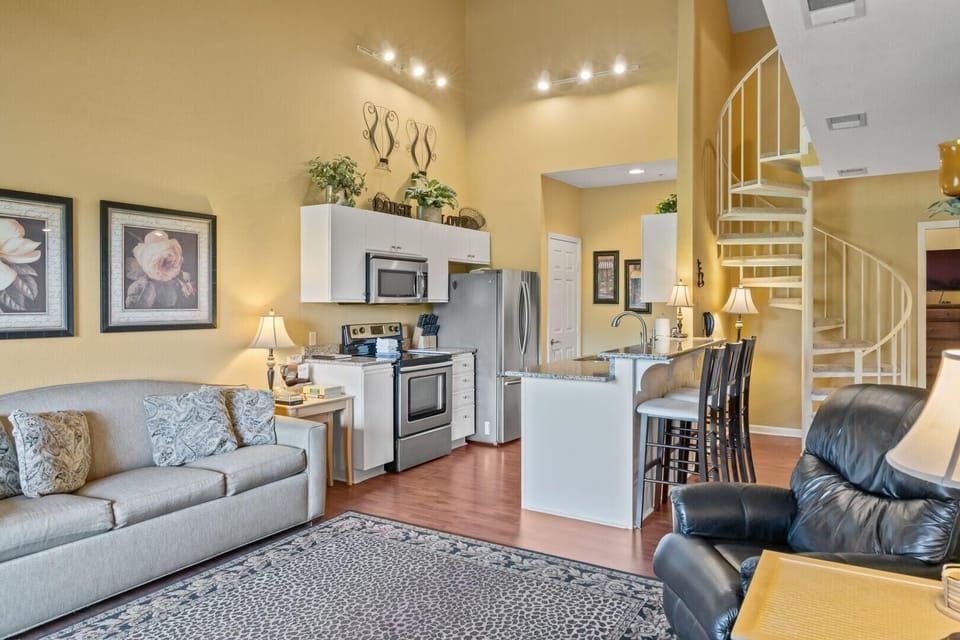 Living area with patterned rug, gray sofa, black leather recliner, and white spiral staircase beside the kitchen bar.