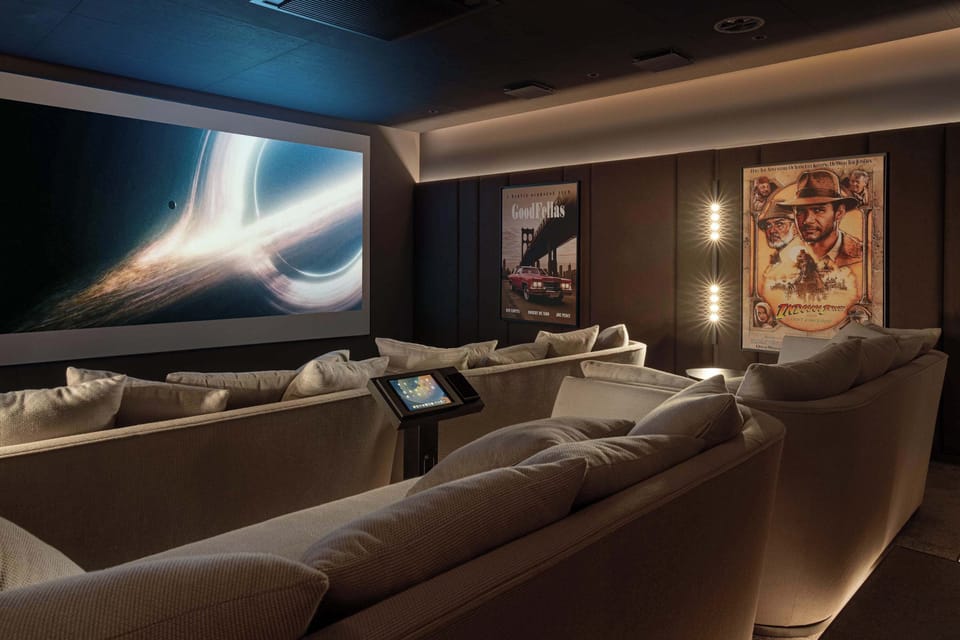 A private home theater room fitted with rows of tiered, comfortable leather recliner seating. A large projection screen displays a movie, flanked by framed cinema posters and soft ambient lighting.