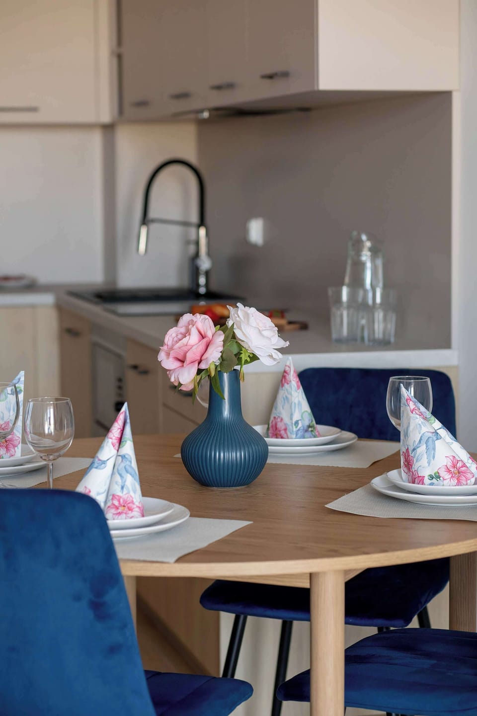 A cozy dining area connected to a compact kitchen, featuring a wooden table set with plates, glasses, and a small decorative vase. Light cabinetry and neutral tones create a warm, inviting space for meals.