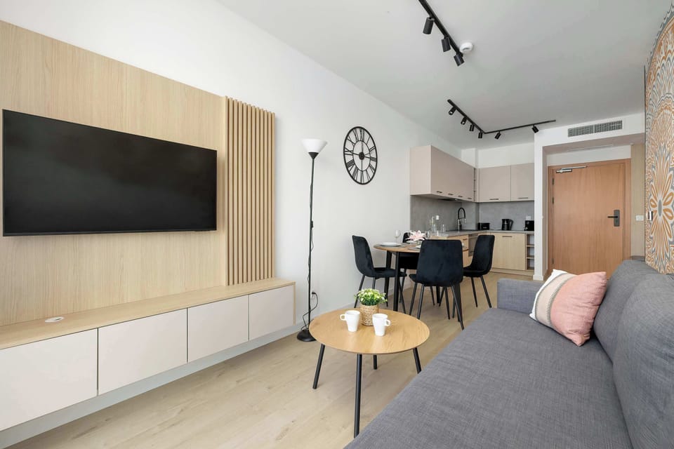 A dark grey sofa sits opposite a wall-mounted TV in a bright living area. The room is decorated with a minimalist aesthetic, featuring neutral tones and wooden accents.