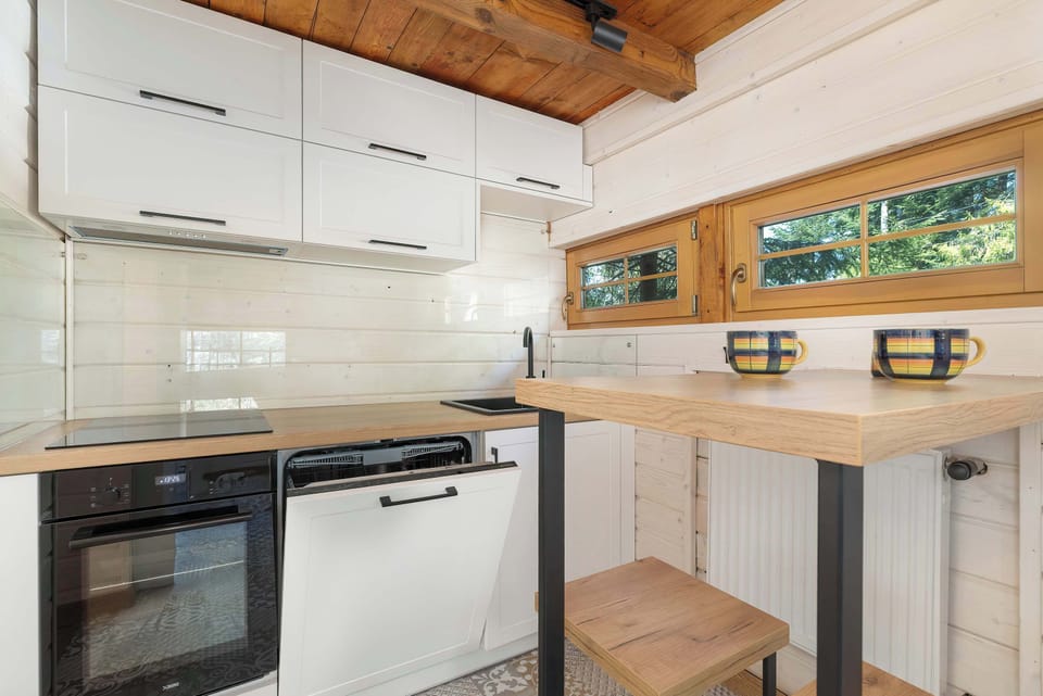 The upper section of the kitchenette displays clean white cupboards and a view of the forest through a small, high window.