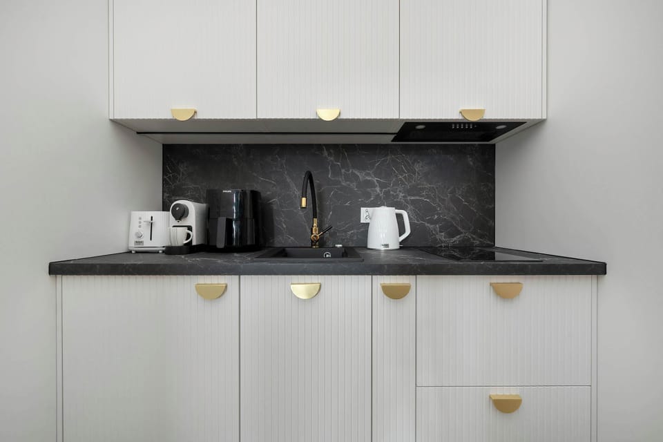 A minimalist kitchenette features sleek white cabinetry with unique gold semi-circle handles and a black marble-effect backsplash. The setup includes an integrated stovetop and a modern white electric kettle.