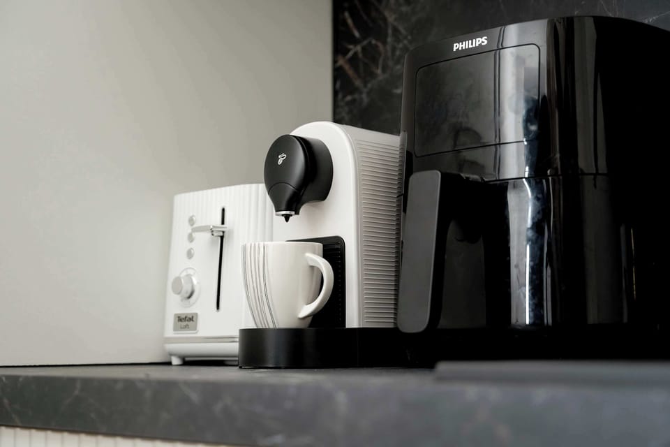 A close-up on the kitchenette counter shows a white capsule coffee machine and a black air fryer. These modern appliances are neatly arranged on a dark, textured worktop.