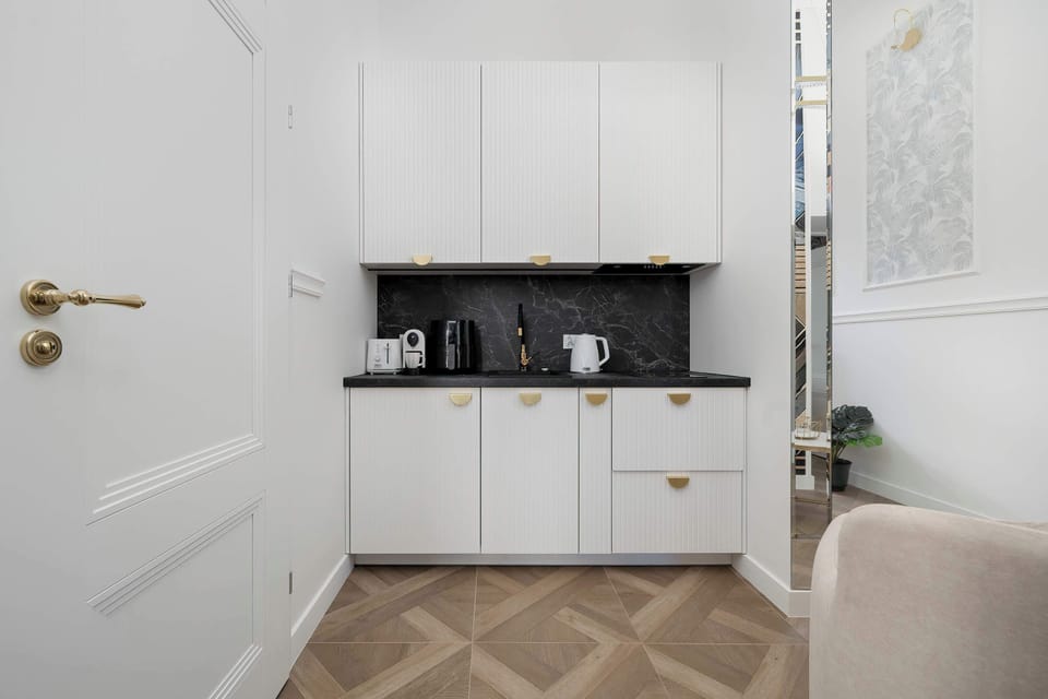 The kitchenette area is shown from the side, featuring minimalist white cabinets and a dark backsplash. The consistent herringbone flooring ties the cooking space into the rest of the apartment.