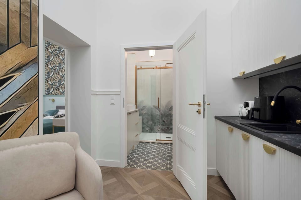 A perspective from the armchair looks through an open white door into the bathroom. The kitchenette area is visible to the right, showing the compact and efficient layout of the apartment.