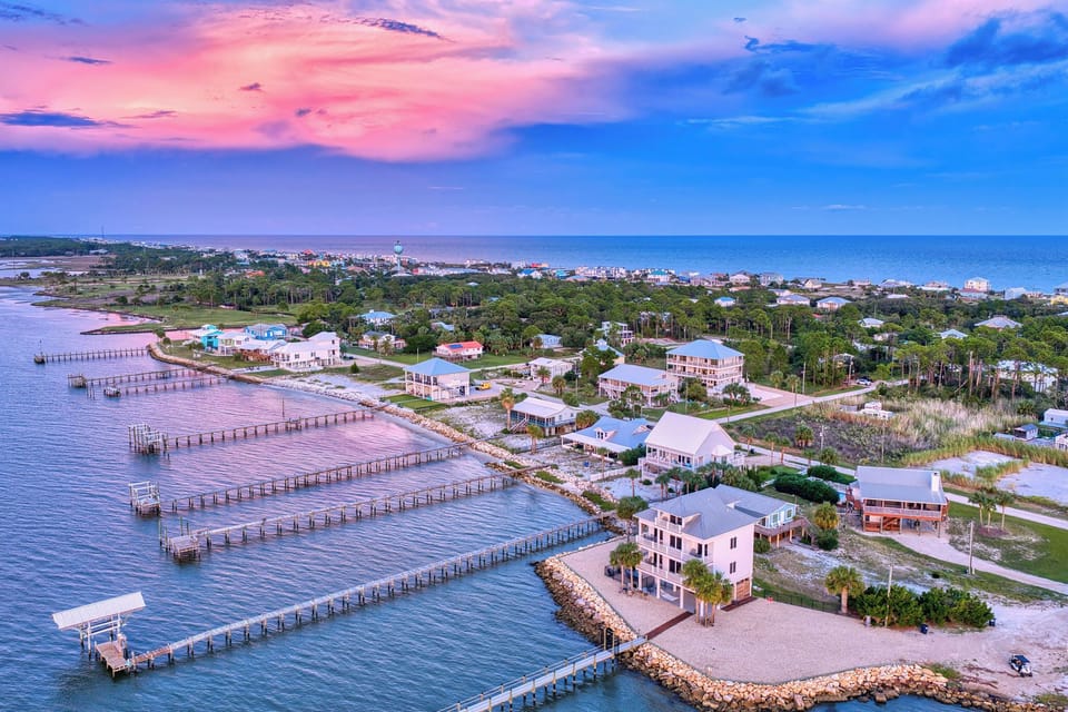 An aerial view of a waterfront community at sunset, with vacation homes nestled among palm trees and long private piers stretching into calm waters.