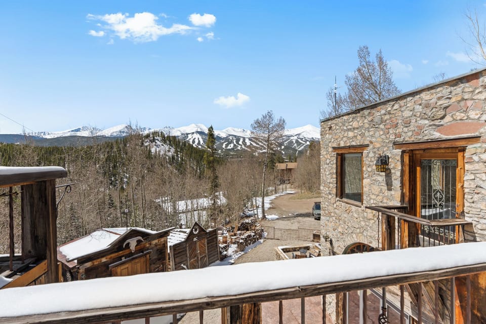 Stunning mountain property with snow-capped peaks visible beyond forested surroundings, featuring rustic stone and wood architecture nestled in a winter landscape.