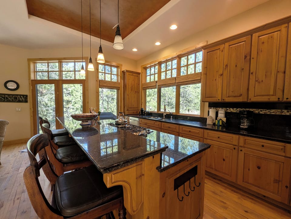 Your gourmet kitchen features granite countertops, warm wood cabinetry, and panoramic forest views through soaring windows.