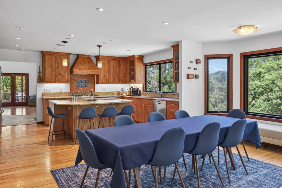 Pendants glow over granite counters while the dining table stretches toward that leafy window bench — pull up a stool for coffee talk.