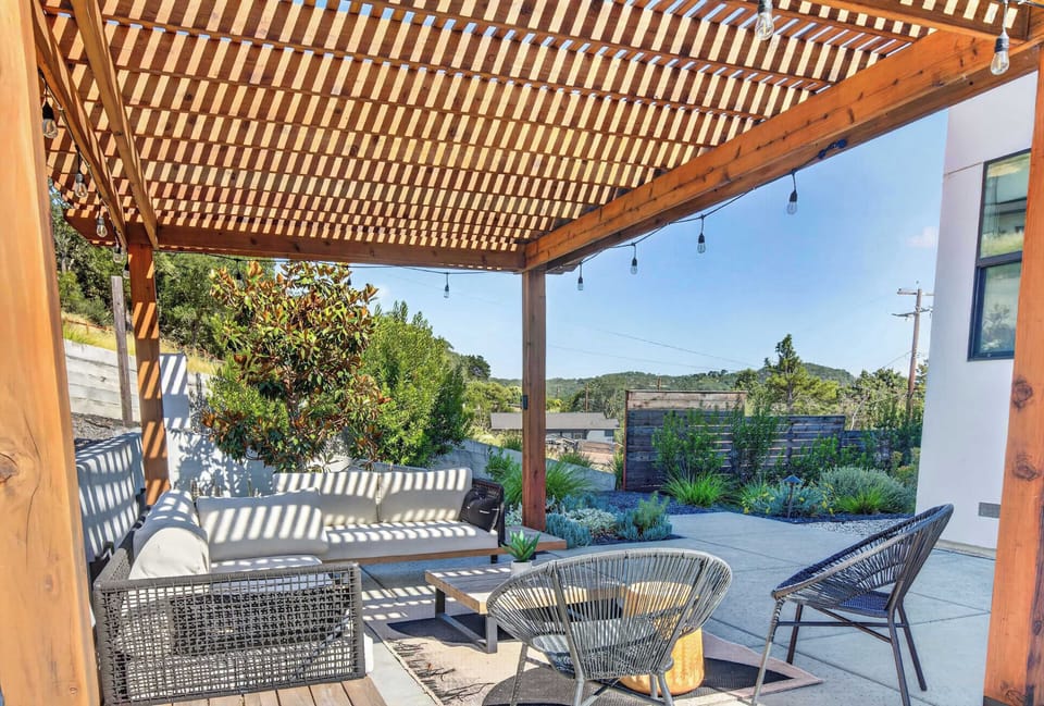 Slatted pergola filters sun over a deep sofa and woven chairs — string lights, succulent beds, and hillside views set the scene.