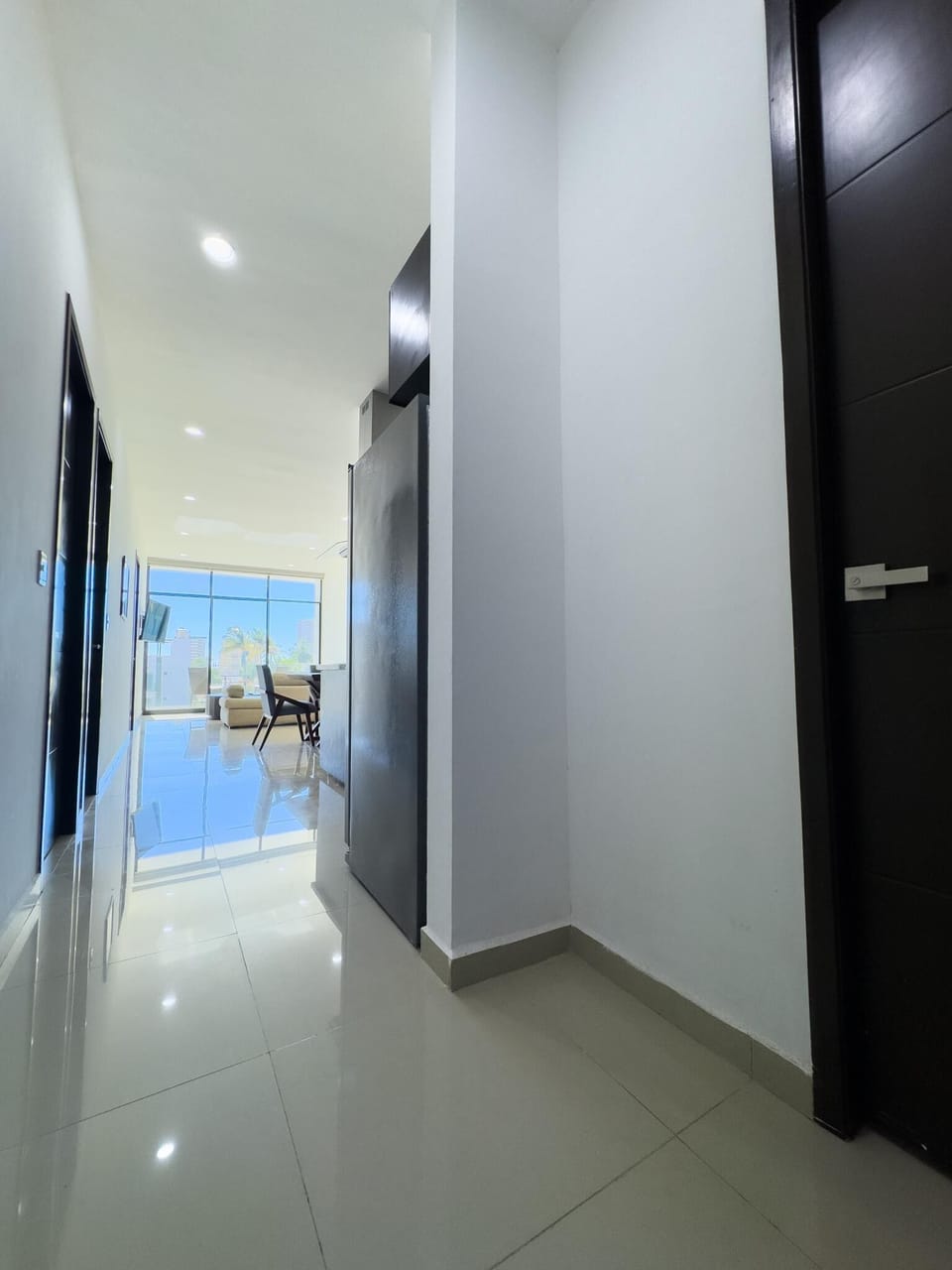 Hallway with glossy cream tiles, white walls, dark wood doors, and recessed ceiling lights leading to living area.