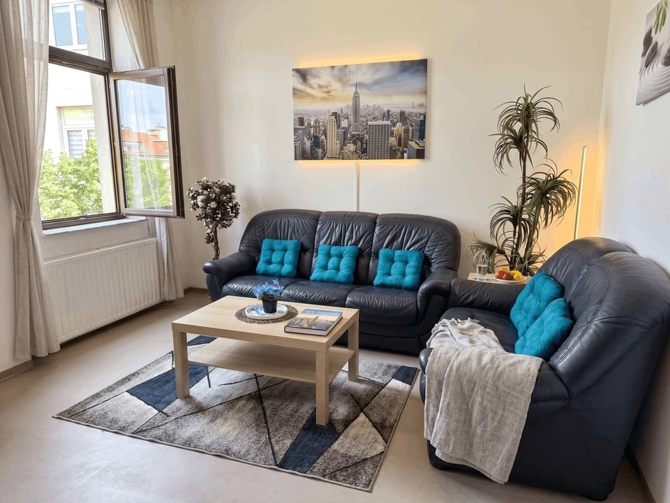 Wohnzimmer – Gemütliches Wohnzimmer mit dunklen Ledersofas, türkisen Kissen, Pflanzen und NYC-Wandbild.