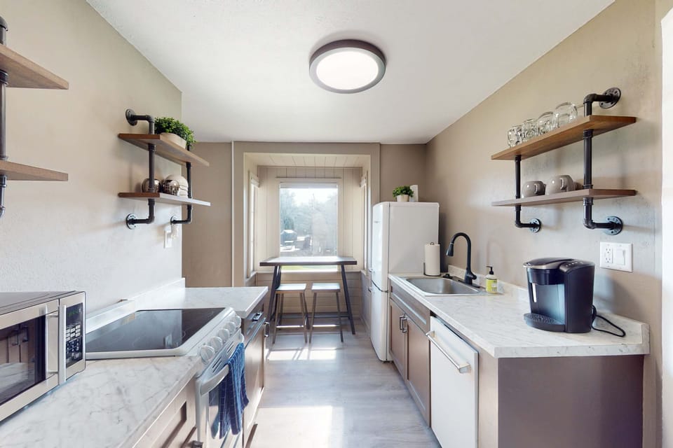 Sunlit galley kitchen with pipe-shelf open storage, Keurig, and electric range — everything you need to skip the restaurant