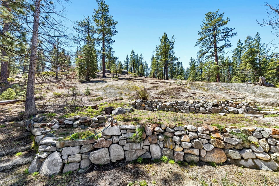 The property sits on a natural hillside with stone terracing and mature pine trees under a bright blue sky, offering a serene mountain setting.