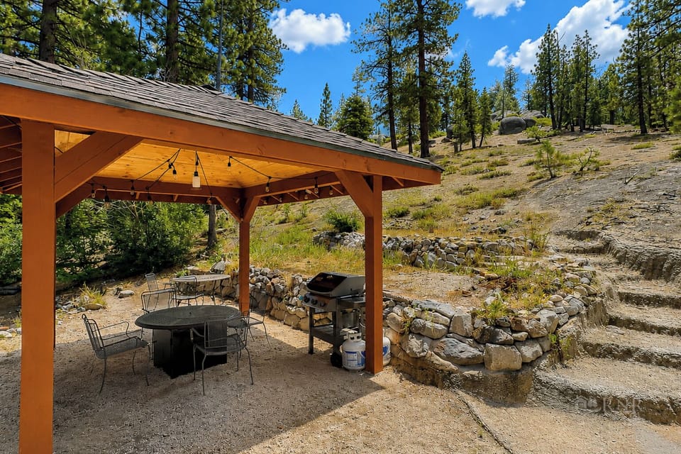 The property features a covered outdoor dining pavilion with BBQ facilities, set against a natural landscape of towering pines and native vegetation under open mountain skies.