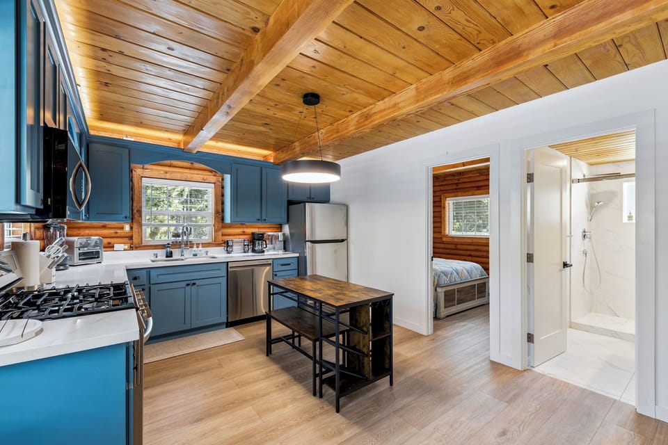 Step into your cozy cabin retreat featuring a fully-equipped kitchen with modern appliances, warm wood-beam ceilings, and stylish blue cabinetry. The open layout flows naturally into the bedroom and bathroom, creating your perfect mountain getaway.