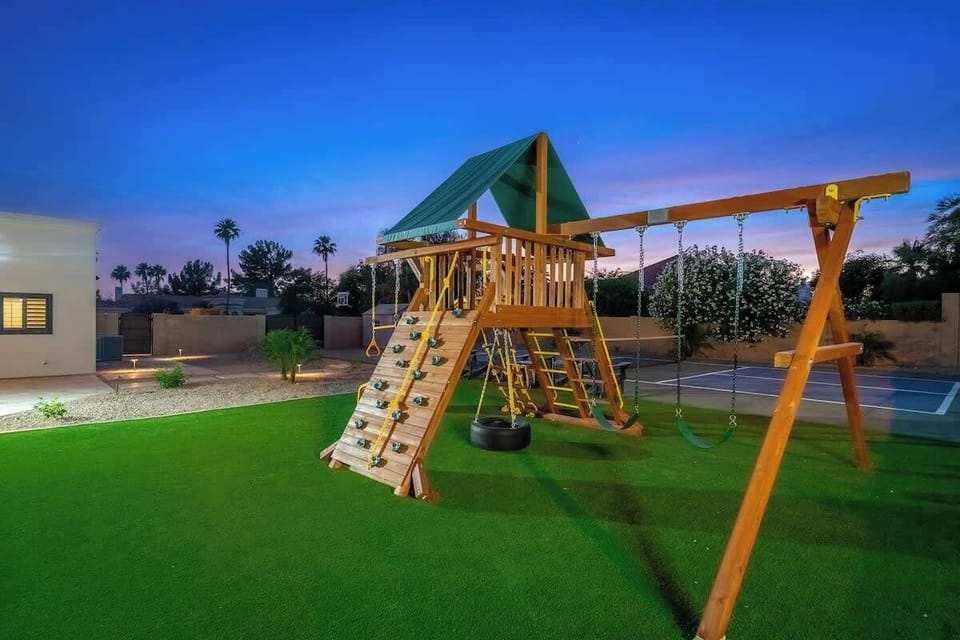 A playground with swings, climbing wall, and covered platform stands ready on lush green lawn, with sports court and palm trees framing the backyard at dusk.