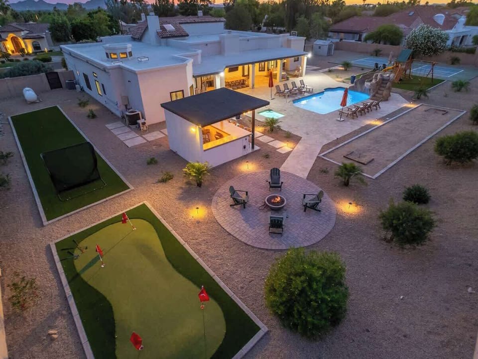 Aerial view of a contemporary vacation home showcasing a sparkling pool, private putting green, fire pit patio, and covered outdoor living areas set against a stunning desert mountain backdrop at sunset.