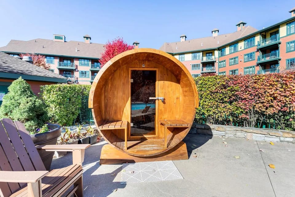 Round wooden outdoor sauna with glass door, built-in benches, and Adirondack chairs on concrete patio.