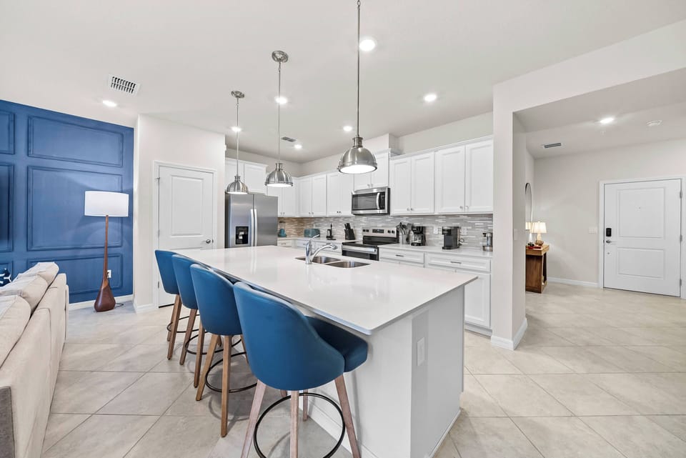 Welcome to a bright kitchen with sleek white cabinetry and a vibrant blue accent wall. The spacious island, paired with stylish bar stools, invites you to gather for fresh meals or morning coffee in a warm, inviting atmosphere.